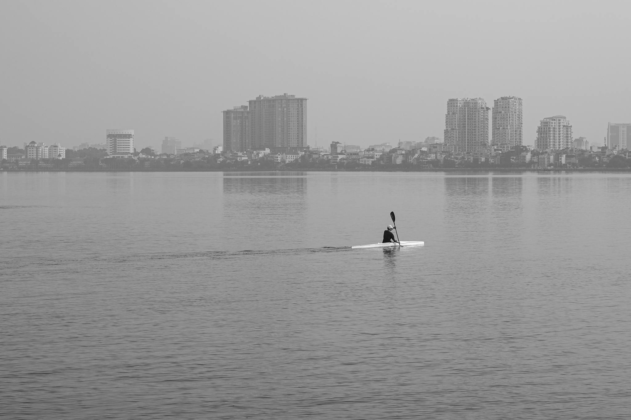 A lone kayaker paddles across a calm urban waterway with city buildings in the background.