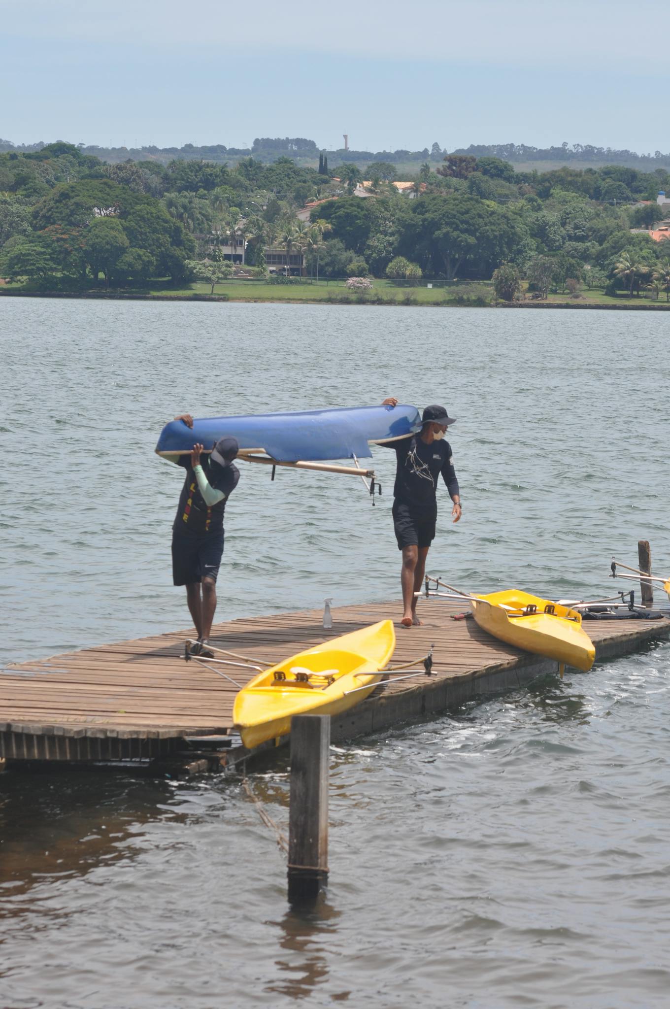 Two men carrying a blue canoe onto a dock with kayaks in Brasília, Brazil.